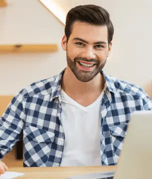 Smiling man with dark hair and a beard, wearing a blue and white checkered shirt over a white t-shirt, sitting at a desk with a laptop in a well-lit modern workspace.