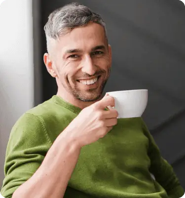 Smiling man with short gray hair wearing a green sweater, holding a white coffee cup while sitting in a modern indoor setting. He appears relaxed and approachable.